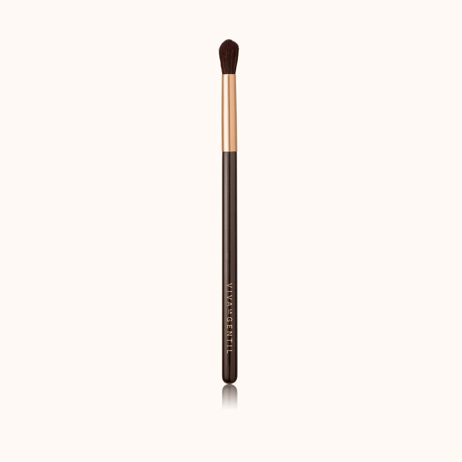 A precise eye crease makeup brush with a rose gold ferrule with a dark brown lacquered handle, made using handcrafted synthetic dark brown bristles mixed with a few strands of white bristles.